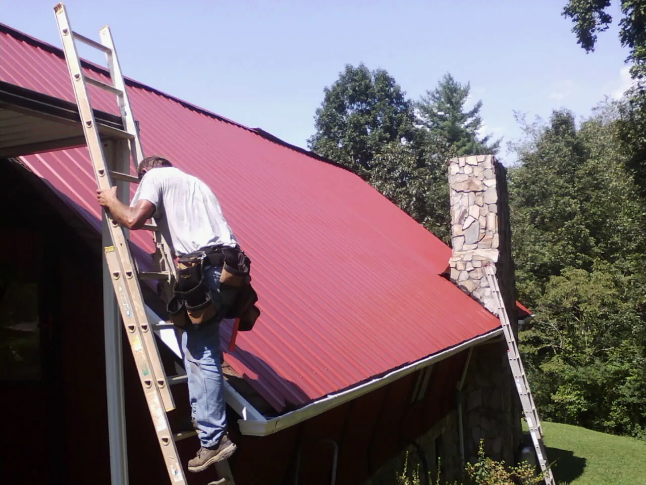 Roofer on a ladder beside a red metal roof and stone chimney — Soffit & Fascia Repair by Roofing Brothers in Upton